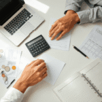 Un spațiu de lucru ordonat, cu un laptop, un calculator, documente financiare, monede, un pix și un registru deschis; mâini care examinează hârtii ...