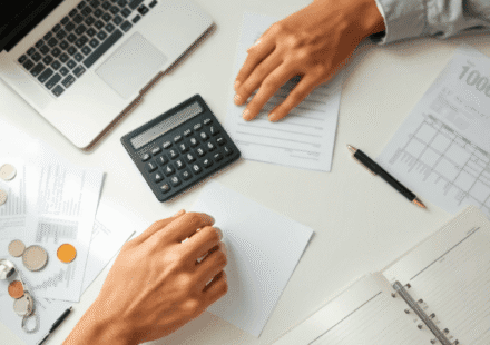 Un spațiu de lucru ordonat, cu un laptop, un calculator, documente financiare, monede, un pix și un registru deschis; mâini care examinează hârtii ...